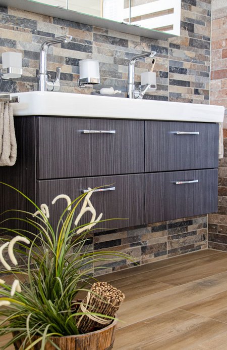 Moderner Doppelwaschtisch mit dunkler Holzfront, zwei Chromarmaturen und gemauerter Wandverkleidung in Natursteinoptik, mit dekorativer Pflanzenaufstellung im Vordergrund.