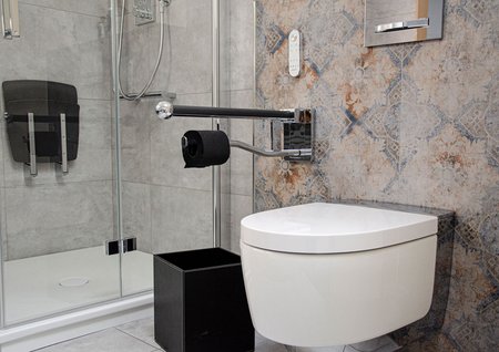 Moderner, barrierearmer Sanitärbereich mit wandhängendem WC, klappbarem Haltegriff, gemusterten Wandfliesen und einer Dusche mit Glasabtrennung.