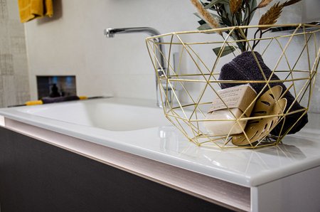 Nahaufnahme eines modernen Waschtischs mit weißer Keramikoberfläche, goldfarbenem Drahtkorb mit Dekoartikeln und Handtuch, in einer hellen Badezimmerausstellung.